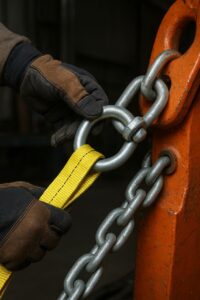 در این تصویر، یک کارگر حرفه‌ای در حال بستن اسلینگ پارچه‌ای زرد به یک حلقه و زنجیر فلزی سنگین است. عملیات اتصال با دستکش ایمنی و تجهیزات استاندارد انجام می‌شود تا بارهای سنگین هنگام جابه‌جایی یا لیفتینگ کاملاً ایمن شوند. این فرایند بخشی از مراحل آماده‌سازی بار برای حمل‌ونقل صنعتی، جرثقیل‌کاری یا مهاربندی تجهیزات سنگین است. استفاده از اسلینگ، شگل و زنجیر مناسب نقش مهمی در ایمنی و کیفیت عملیات جابه‌جایی دارد.